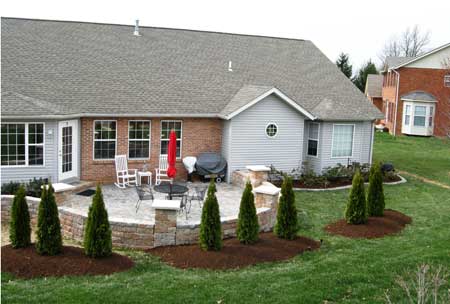 Elevated view of rear showing stone patio, grass, mulch beds and tree lines